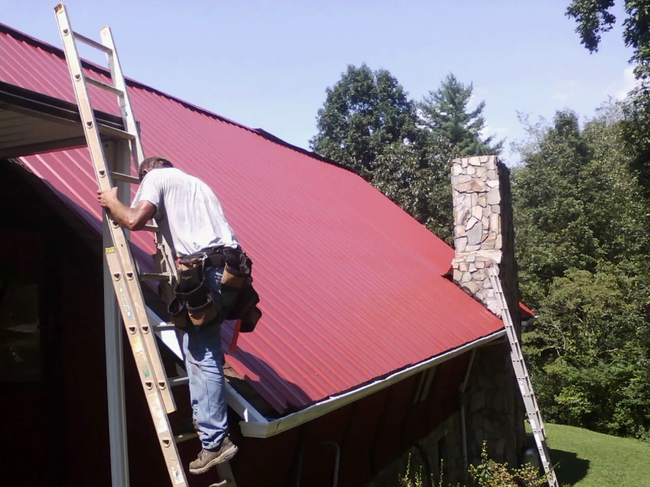 Roofer on a ladder beside a red metal roof and stone chimney — Soffit & Fascia Repair by Roofing Brothers in Elmaton